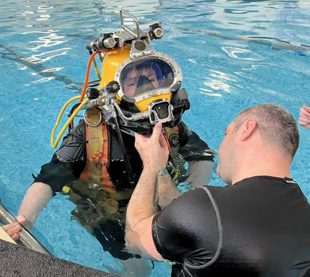 An intro to Commercial Diving A man helping a boy in a pool with a diving helmet.