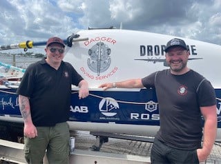 Two men smiling next to a boat on land.